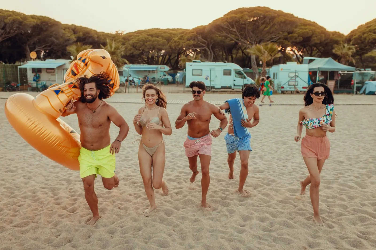 Vrienden rennen over het zand naar zee, met de camping en parasoldennen vlak achter het strand